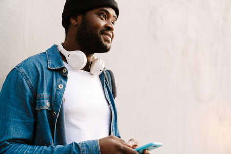Black young man with headphones smiling and using mobile phone outdoorsの写真素材