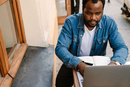Black young man working with laptop while sitting in cafe outdoors