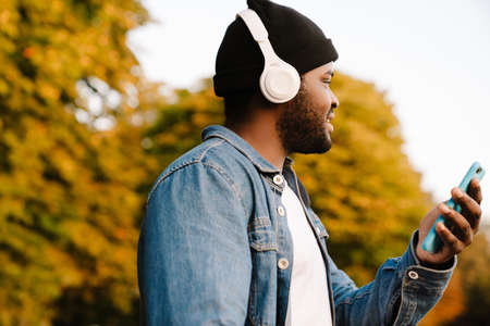 Black man in headphones using mobile phone while walking in autumn parkの写真素材