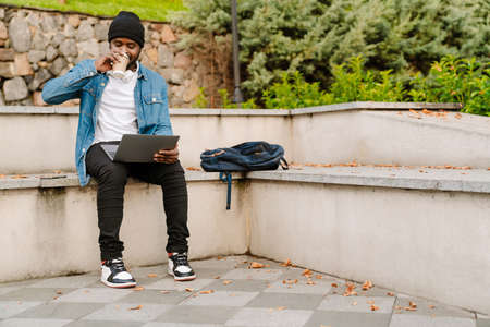Black young man working with laptop while drinking coffee on bench in parkの写真素材