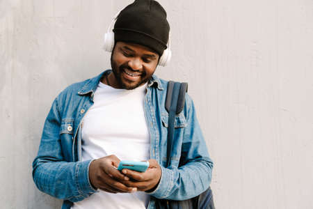 Black young man with headphones smiling and using mobile phone outdoors