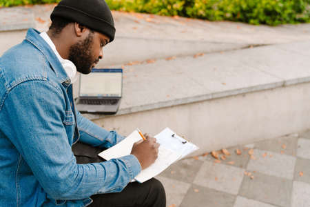 Black man working with laptop and papers while sitting on bench in park