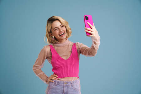 Young white woman smiling while taking selfie on cellphone isolated over blue wallの写真素材