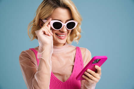 Young white woman in sunglasses smiling while using mobile phone isolated over blue wall