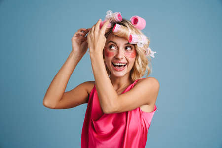 Young woman with curlers and eye patches smiling at camera isolated over blue wallの写真素材
