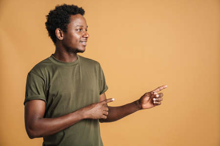 Young black man wearing t-shirt smiling and pointing fingers aside isolated over beige background