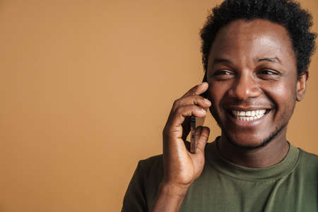 Young black man wearing t-shirt laughing and talking on cellphone isolated over beige backgroundの写真素材