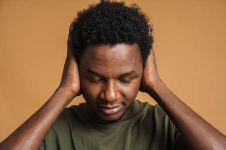 Young black man wearing t-shirt posing while covering his ears isolated over beige background