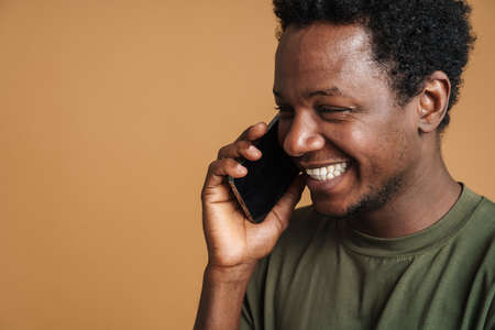 Young black man wearing t-shirt laughing and talking on cellphone isolated over beige backgroundの写真素材