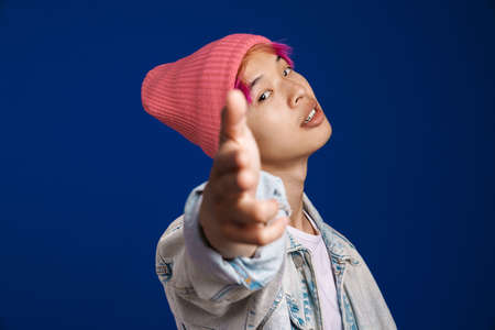 Asian boy in pink hat looking and pointing fingers at camera isolated over blue backgroundの写真素材