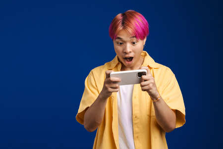 Asian boy with pink hair playing video game on cellphone isolated over blue backgroundの写真素材