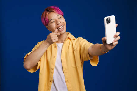Asian boy with pink hair gesturing while taking selfie on cellphone isolated over blue backgroundの写真素材