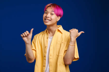 Asian boy with pink hair laughing and pointing fingers aside isolated over blue backgroundの写真素材