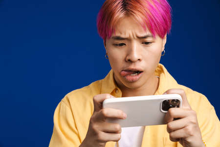 Asian boy with pink hair playing online game on mobile phone isolated over blue backgroundの写真素材