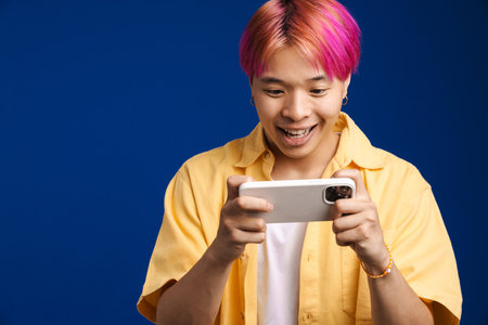 Asian excited boy with pink hair playing online game on mobile phone isolated over blue backgroundの写真素材