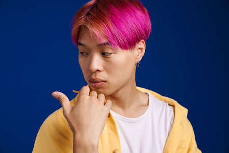 Asian boy with pink hair looking and pointing finger at camera isolated over blue backgroundの写真素材