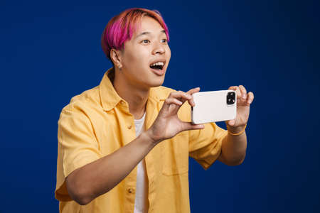 Asian boy with pink hair smiling while taking photo on mobile phone isolated over blue backgroundの写真素材