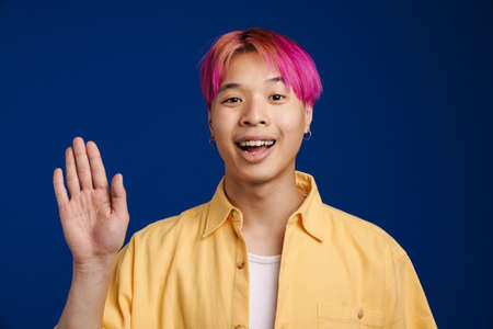 Asian boy with pink hair smiling while waving hand isolated over blue backgroundの写真素材