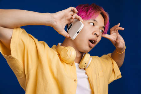 Asian boy pointing finger at his temple while talking on cellphone isolated over blue backgroundの写真素材
