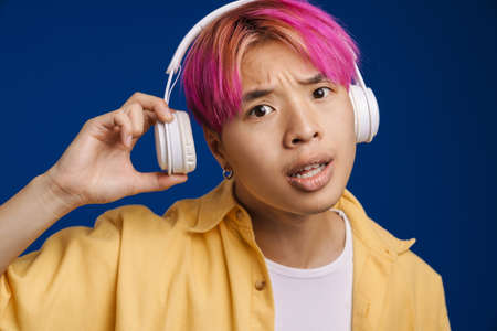 Asian boy with pink hair listening music with headphones isolated over blue background