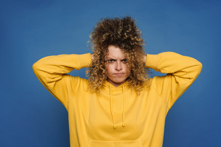 Blonde woman dressed in hoodie frowning and holding her head isolated over blue backgroundの写真素材