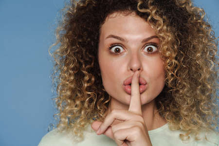Young blonde woman expressing surprise and showing silence gesture isolated over blue backgroundの写真素材