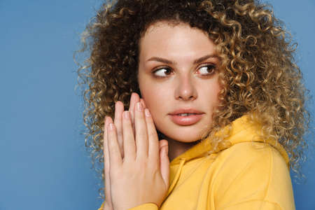 Young blonde woman in hodie holding palms together and looking aside isolated over blue background