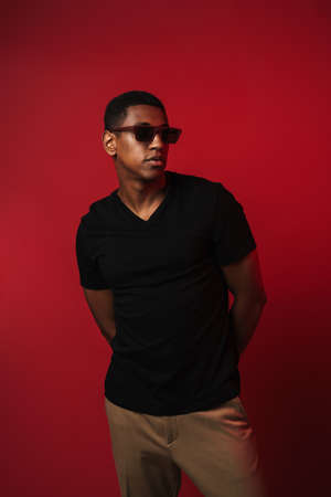 Young black man wearing sunglasses posing and looking aside isolated over red wall