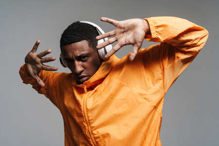 Young black man gesturing while listening music with headphones isolated over grey wall