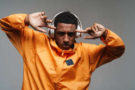Young black man in wireless headphones pointing fingers at his temples isolated over grey wall