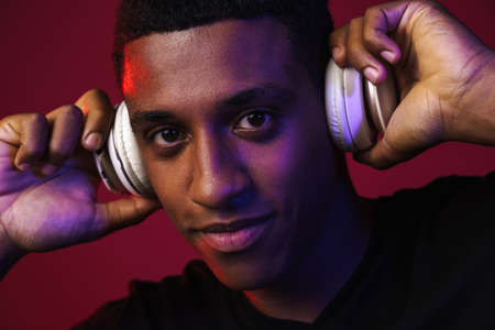 Young black man smiling while listening music with headphones isolated over red wallの写真素材