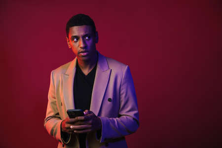 Young black man dressed in jacket using mobile phone isolated over red wallの写真素材