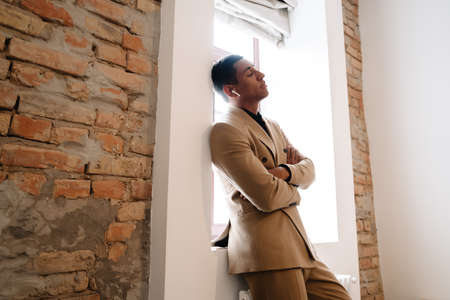 Black man wearing suit using wireless earphones while standing by window indoorsの写真素材
