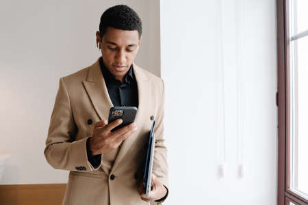 Black man wearing suit using cellphone and wireless earphones indoors