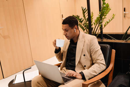 Black man in earphones drinking coffee while working with laptop at hotel