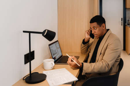 Black man talking on cellphone while working with laptop at desk in hotel