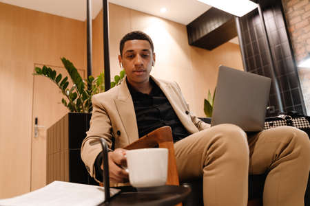 Black man in earphones drinking coffee while working with laptop at hotel