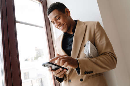 Black man wearing suit using cellphone while standing by window indoorsの写真素材