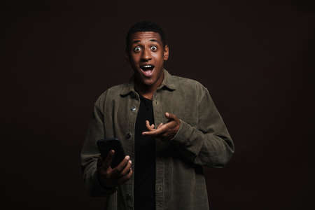 Shocked black man pointing finger aside while using cellphone isolated over brown wall