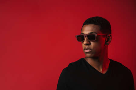 Young black man wearing sunglasses posing and looking aside isolated over red wall