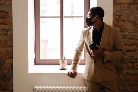 Black man in face mask using wireless earphones and cellphone by window indoors