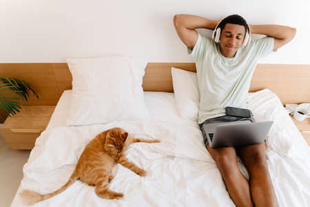 Black man in headphones using laptop while sitting with his cat on bad in hotelの写真素材