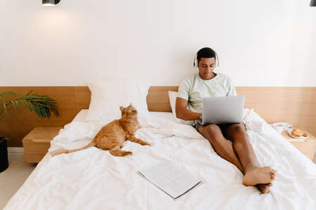 Black man in headphones using laptop while sitting with his cat on bad in hotelの写真素材