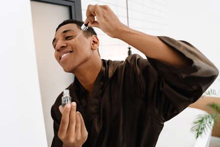 Black man wearing bathrobe applying face serum at morning in bathroom