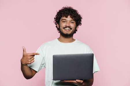 Young middle eastern man smiling while pointing finger at his laptop isolated over pink wall
