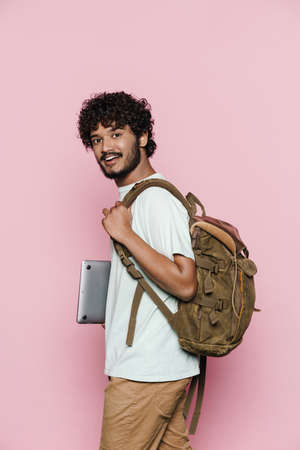 Young middle eastern man smiling while posing with laptop and backpack isolated over pink wall