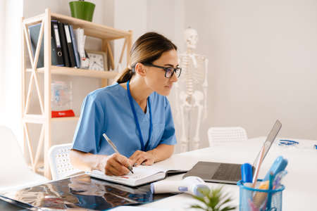 White woman doctor writing down notes while working with laptop in office indoorsの写真素材