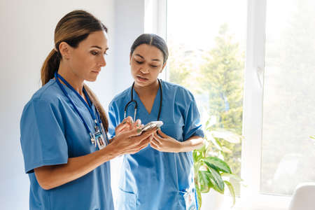 Multiracial women doctors talking and using cellphone while working in office indoors