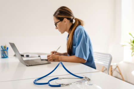 White woman doctor writing down notes while working with laptop in office indoorsの写真素材