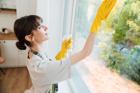 White young woman in gloves smiling while cleaning window at homeの写真素材